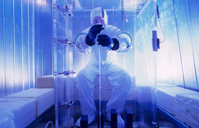 man in white cleanroom suit with hood, mask, and gloves sitting on a white parallelepiped operating on a long thin transparent tube