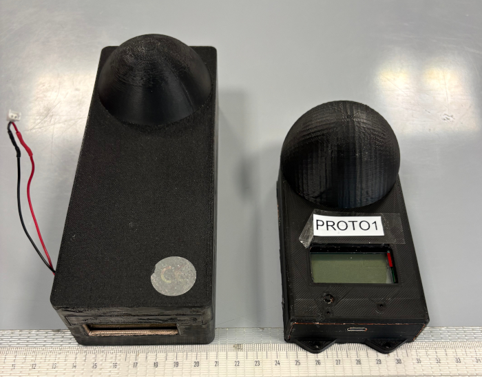 Image of two DOIN dosimeters: rectangular black plastic objects with a hemisphere protruding from one of the large faces. On the left is the first version, which is larger; on the right is a smaller optimized version equipped with a display similar to that of a calculator.