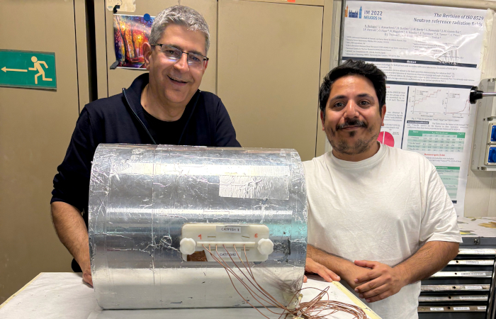 Two smiling people behind a cylinder-shaped neutron spectrometer covered in aluminum. The person on the left has glasses and is wearing a black t-shirt; the one on the right has a mustache and is wearing a white t-shirt. The spectrometer is approximately 50 centimeters large.