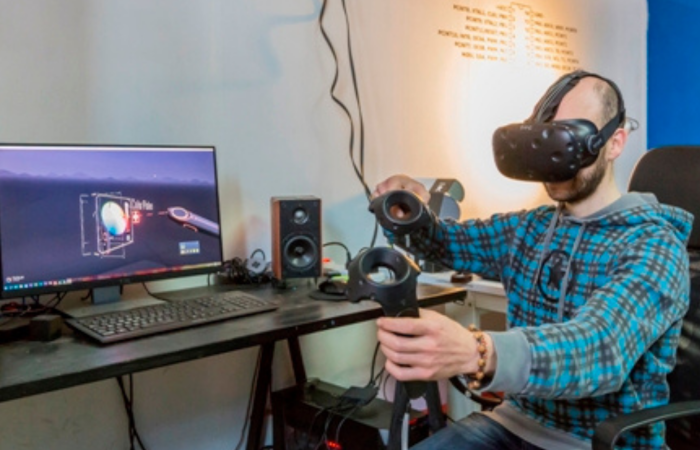 persona seduta a una scrivania di fronte a un monitor, che indossa visore per realtà virtuale e tene in mano dei dispositivi di controllo
