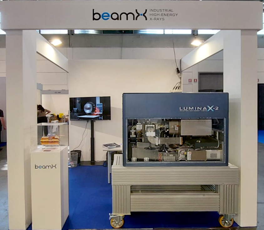 stand in fiera con il logo beamX e l'acceleratore compatto Lumina X2 in esposizione. Lumina X2 è un parallelepipedo largo circa 2 metri e alto circa uno di colore blu, un pannello trasparente mostra l'interno